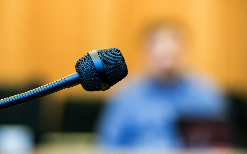 Ein unscharf fotografierter Zeuge im Gerichtssaal hinter dem Mikrofon
